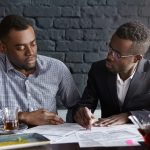 CEO in elegant suit and glasses pointing pen at papers on table in front of him, asking his financial expert to explain figures in report, staring at him with interrogative look. People and business